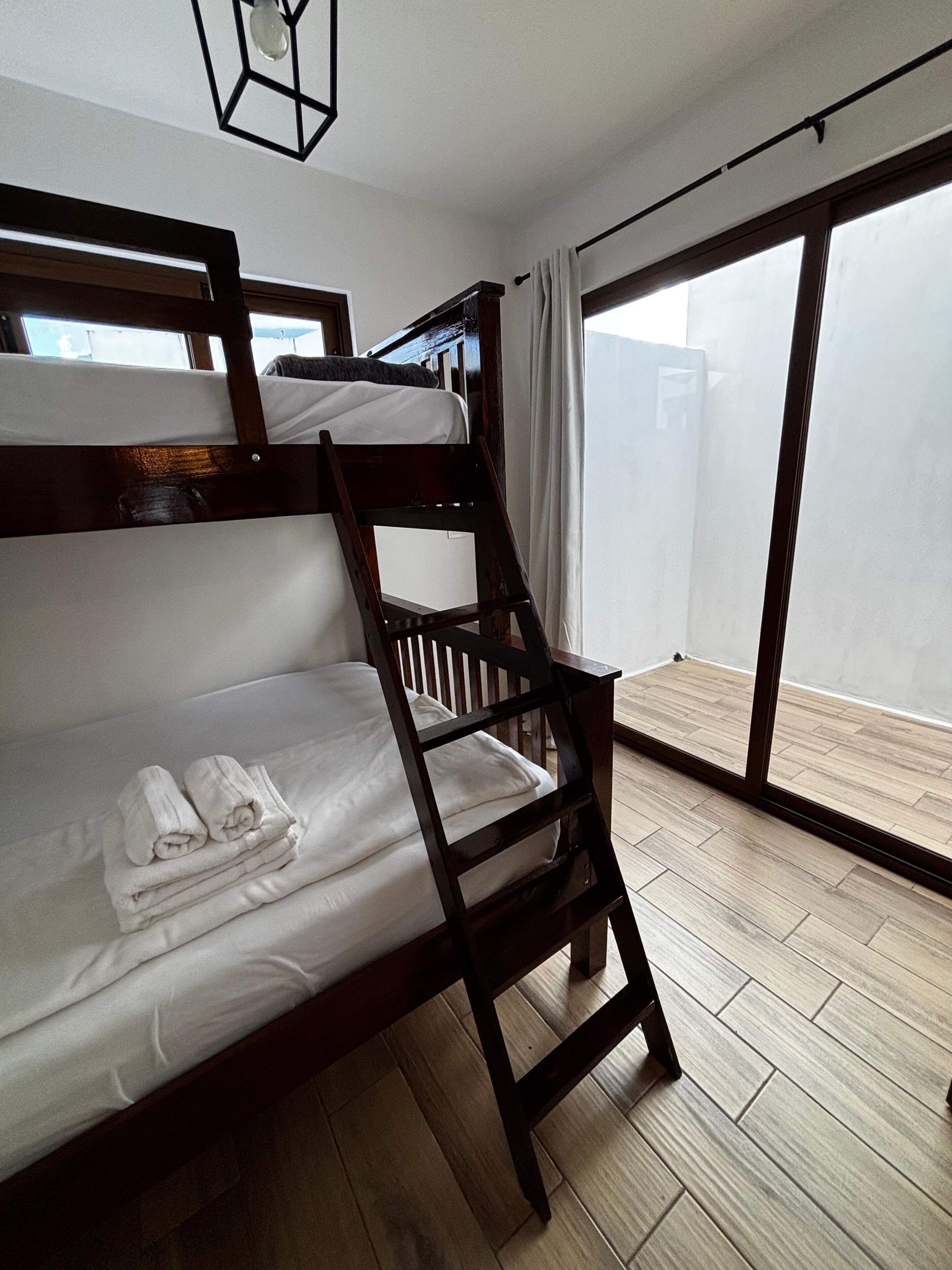 A bunk bed with white bedding and towels, a wooden floor, and a large window with curtains.
