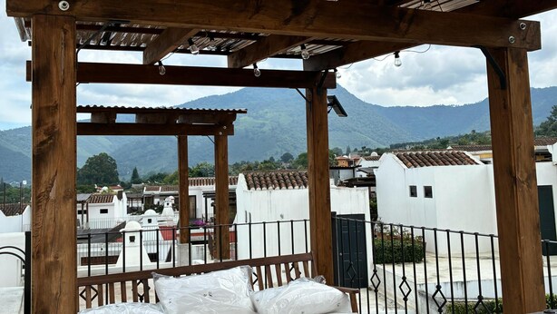 Une pergola en bois, surmontée d’un toit en métal, offre une vue sur un quartier résidentiel avec des bâtiments blancs et des montagnes en arrière-plan.