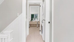 Un pasillo estrecho con una lavadora, una ventana con persianas y una puerta que da a una habitación.