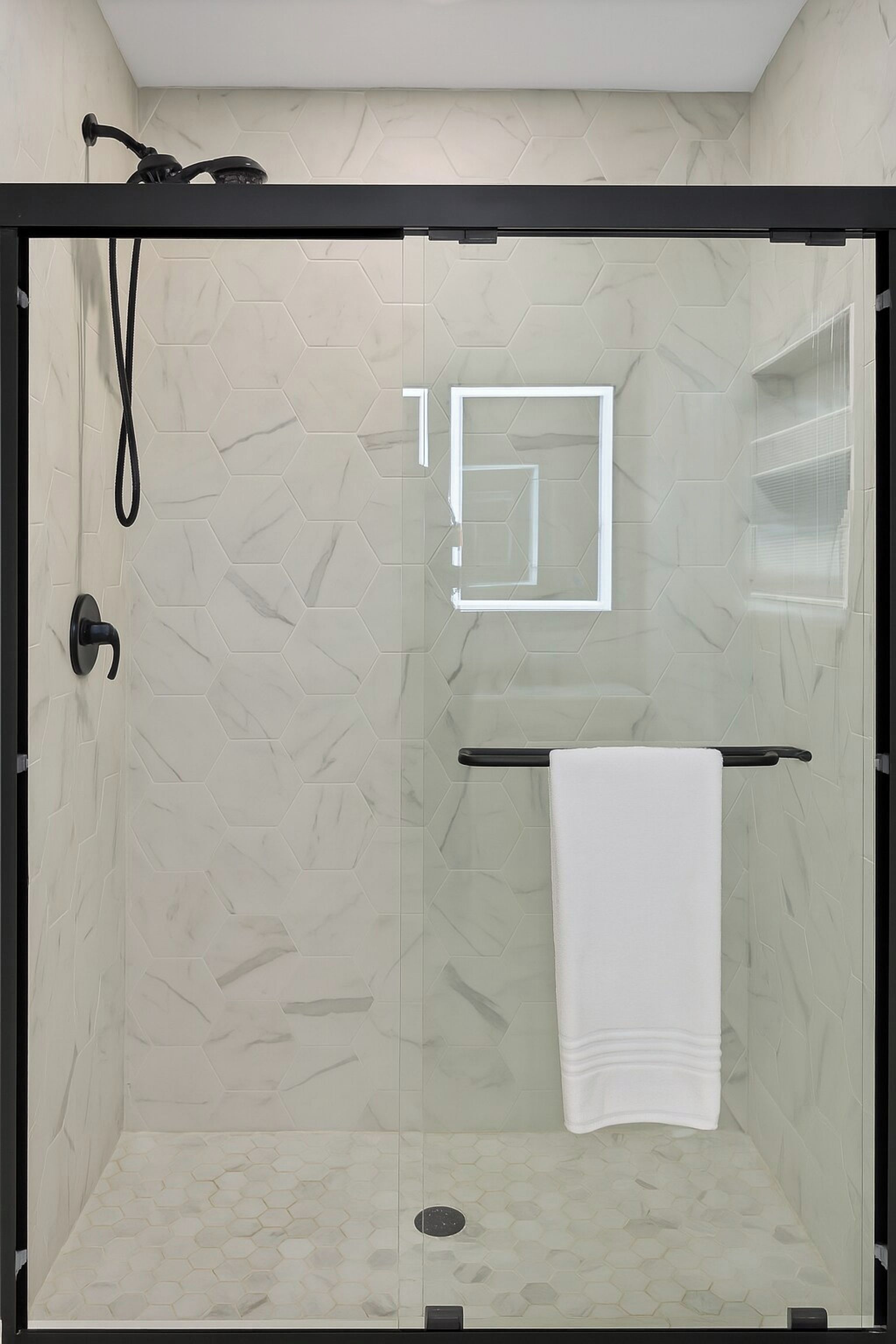Box de vidro com toalha branca em barra preta, azulejos com padrão hexagonal e chuveiro visível.
