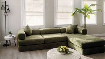 A sectional sofa with a side table and a potted plant in a room with large windows and a floor lamp.