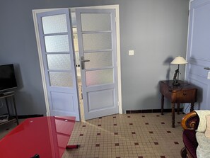 A room with a tiled floor, a closed door with frosted glass panels, a small table with a lamp, and a television on a stand. - A stopover in the heart of the Basque Country (Halsou)