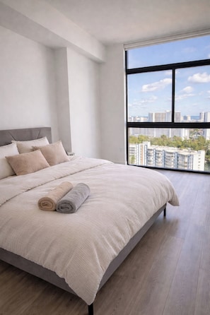 A modern bedroom with a large bed, a view of the city through a window, and a cityscape visible outside. - Large 3BR / pool / gym / Jacuzzi / Sauna / grill (Hallandale Beach)