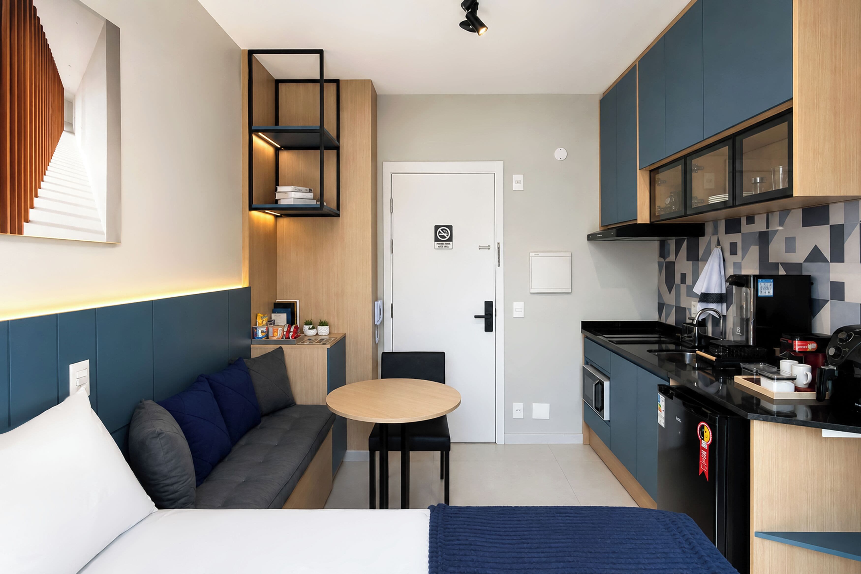 Quarto de hotel compacto com cama, sofá, pequena mesa de jantar, cozinha compacta e banheiro.