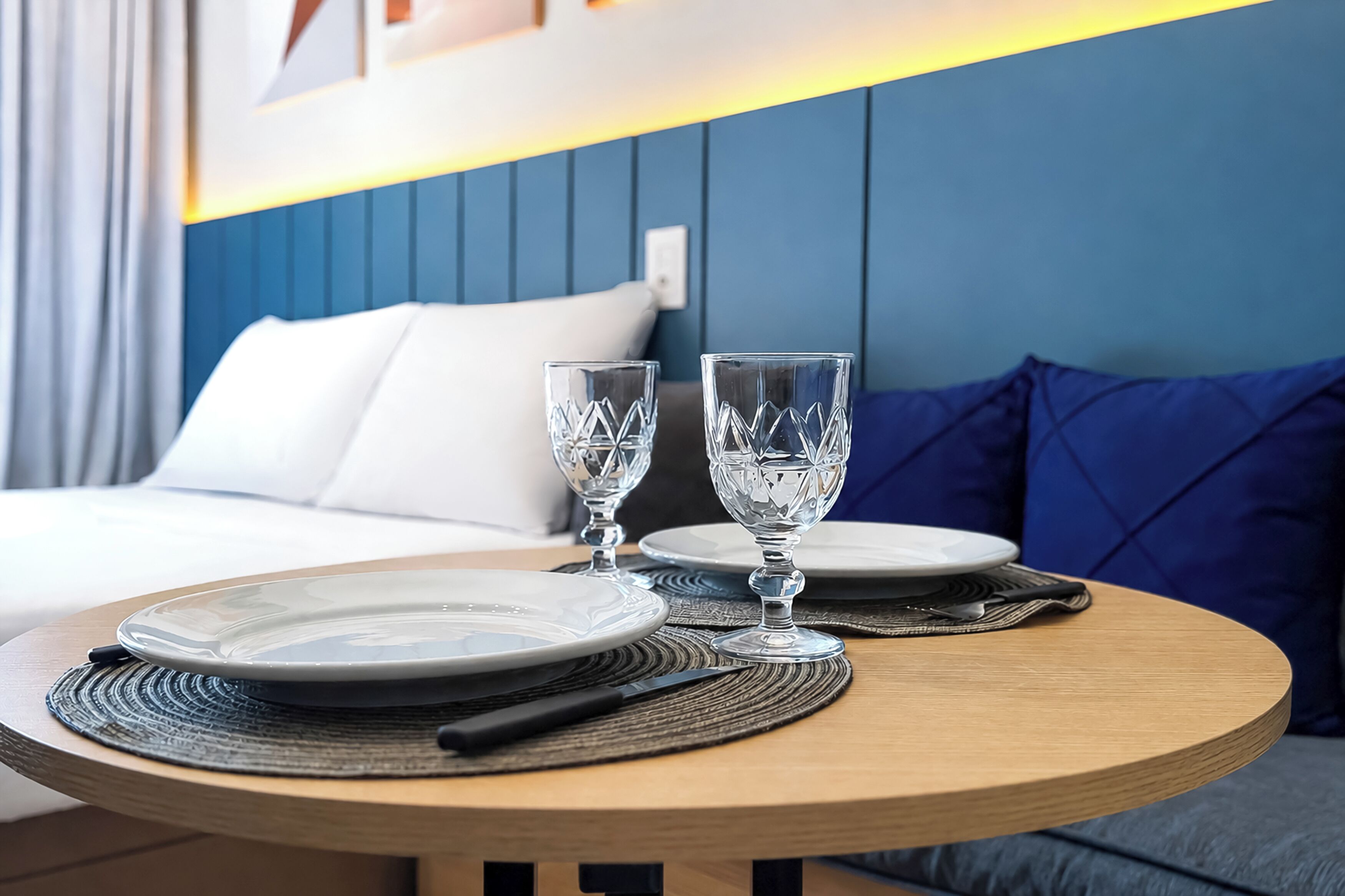 A neatly arranged dining table with plates and glasses, set against a blue wall with a bed in the background.