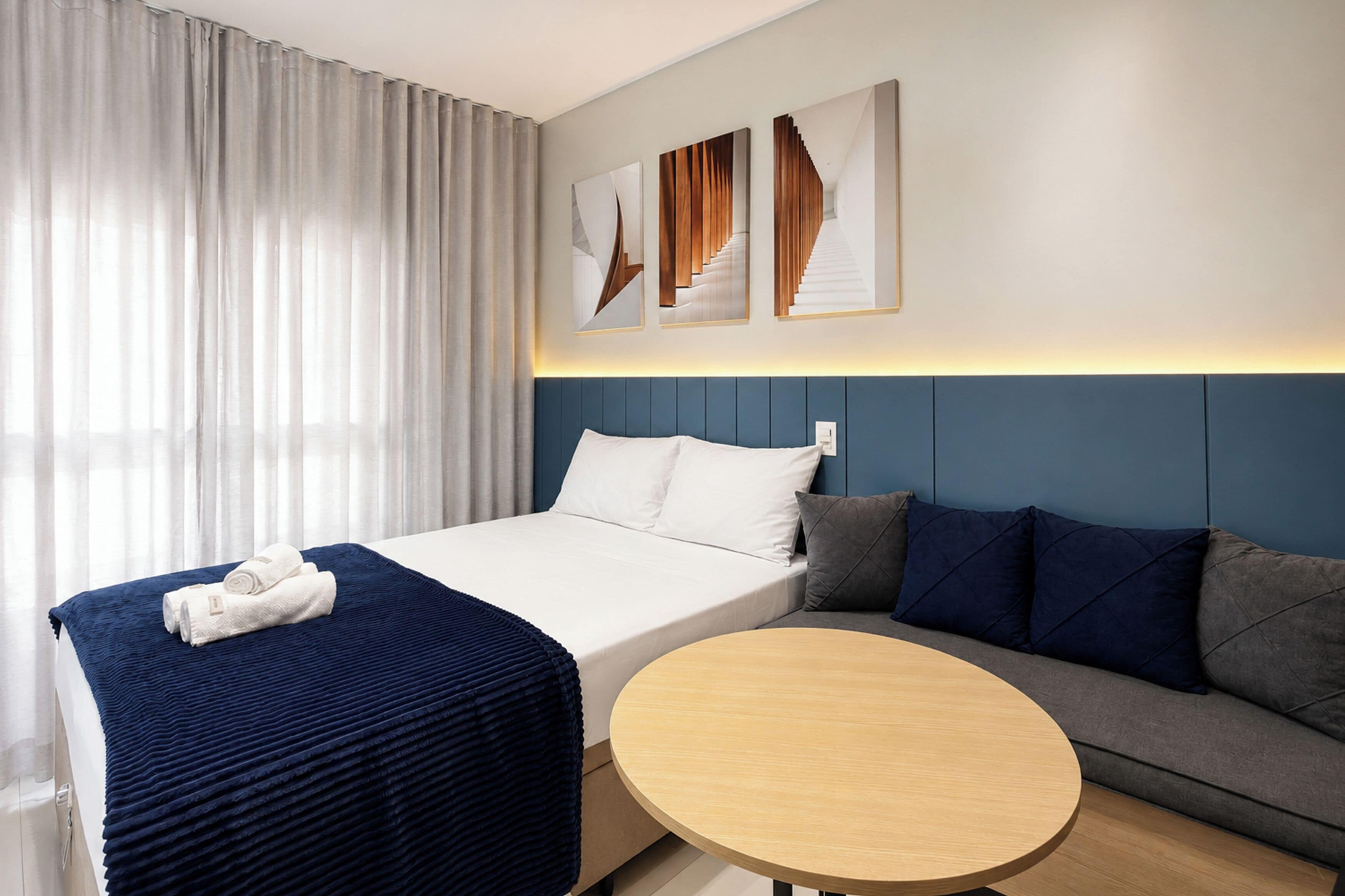 A hotel room with a bed, a round table, a sofa, and a large window with curtains.