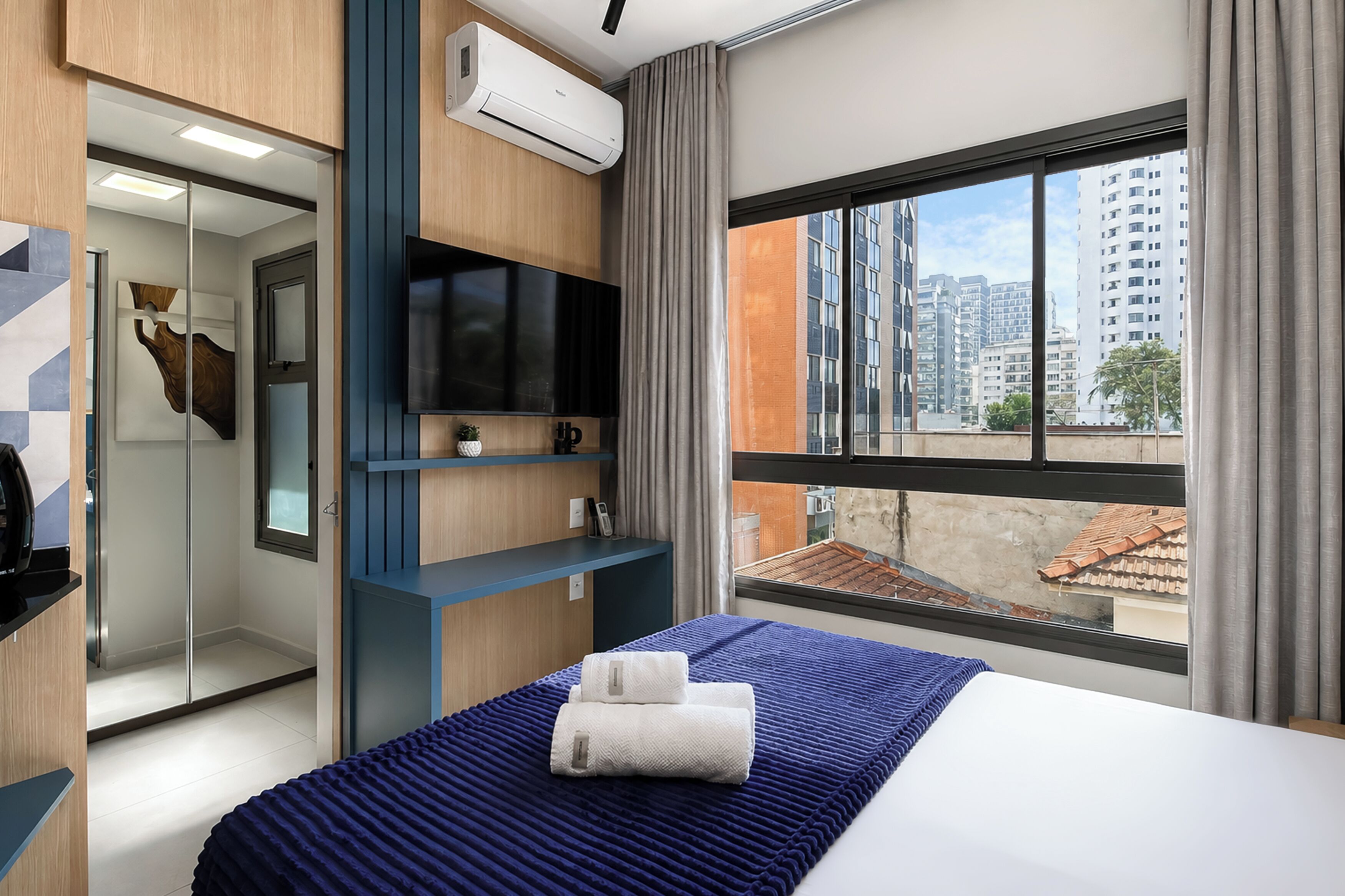 Ein modernes Hotelzimmer mit einem großen Bett, einem Flachbildfernseher, einer integrierten Klimaanlage und Blick auf die Stadtlandschaft.