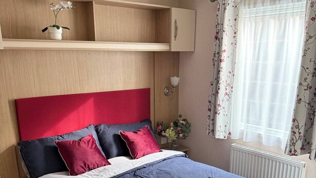 Ein Schlafzimmer mit Bett, Nachttisch, einer Wandablage mit Topfpflanze, einem Fenster mit floralem Vorhang und einem Heizkörper.