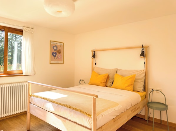 Ein Schlafzimmer mit einem hölzernen Bett, einem Heizkörper, einem Fenster mit Vorhängen, einer Wandlampe und einem gerahmten Bild.