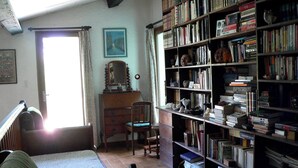 Ein Zimmer mit einem großen Bücherregal, einem Sessel, einem Schminktisch und einem Fenster mit Vorhängen.