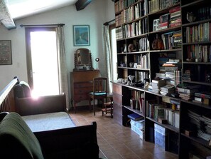 Ein Zimmer mit einem großen Bücherregal, einem Sessel, einem Schminktisch und einem Fenster mit Vorhängen.