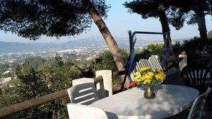 Eine Terrasse mit Tisch und Stühlen, einer Vase mit gelben Blumen und Blick auf eine Stadt in der Ferne.