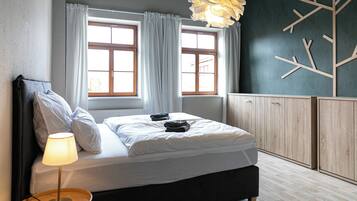 Ein Schlafzimmer mit einem Bett, einem Nachttisch, einer Lampe, zwei Fenstern und einer Wand mit einem geometrischen Muster.