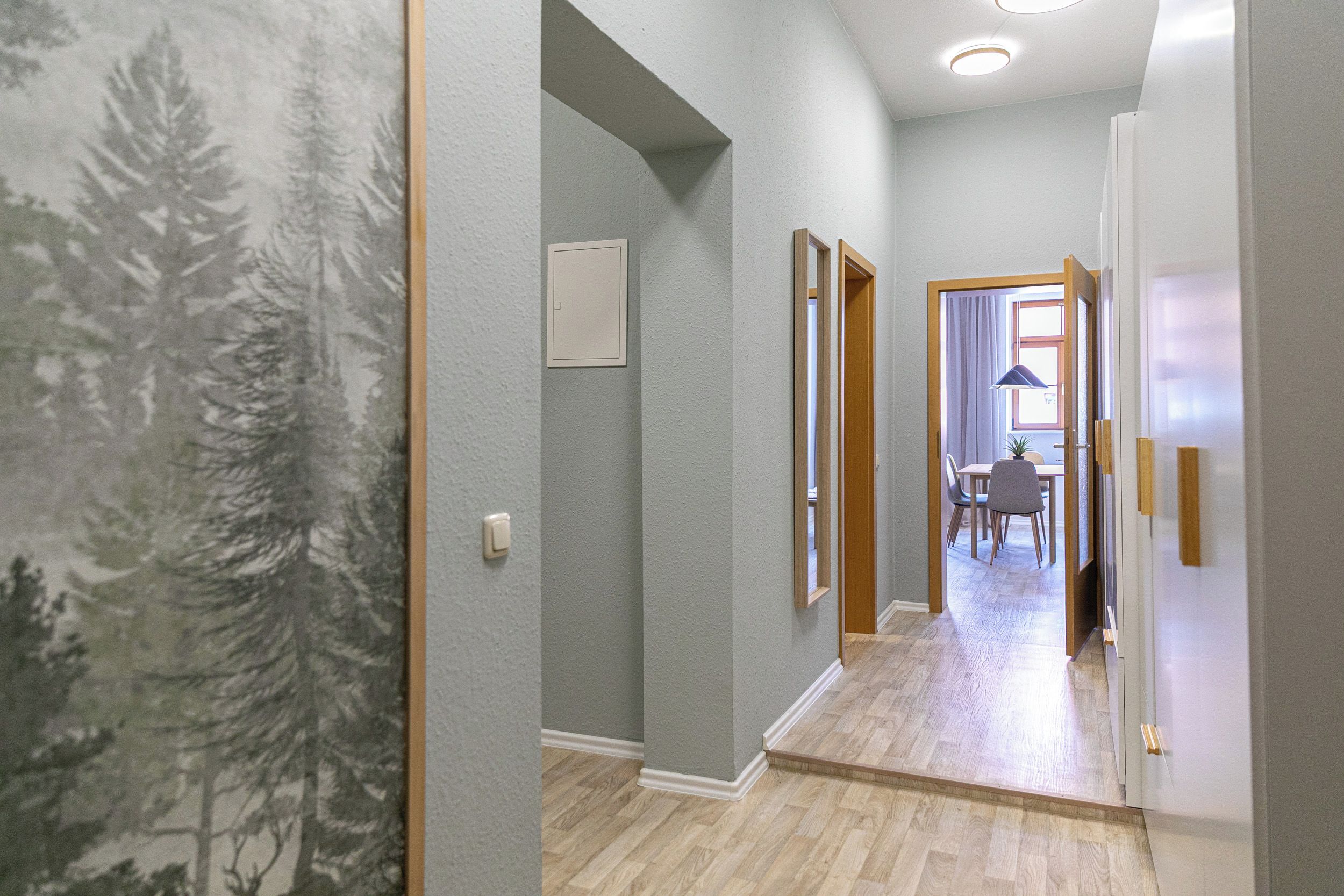 Ein Flur mit einer hellgrauen Wand, einem Holzboden und einer Tür, die in einen Essbereich mit Tisch und Stühlen führt.
