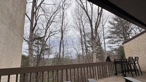 Um deck de madeira com cadeiras pretas e uma mesa, com vista para uma área arborizada.