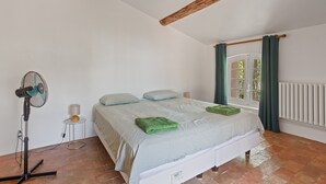 Ein Schlafzimmer mit Bett, Standventilator, Heizkörper und einem fenster mit Vorhängen.