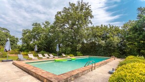 Ein rechteckiger Pool mit Backsteinrand, umgeben von Liegestühlen und Sonnenschirmen, mit Bäumen im Hintergrund.