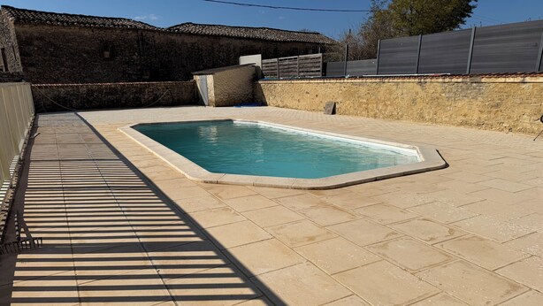 Una piscina rectangular amb un solàrium revestit de rajoles, envoltada per murs de pedra i una tanca de fusta.
