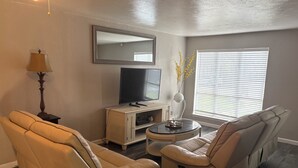 Ein Wohnzimmer mit einer Couch, einem Liegesessel, einem Fernseher, einer Lampe und einem Fenster mit Jalousien.