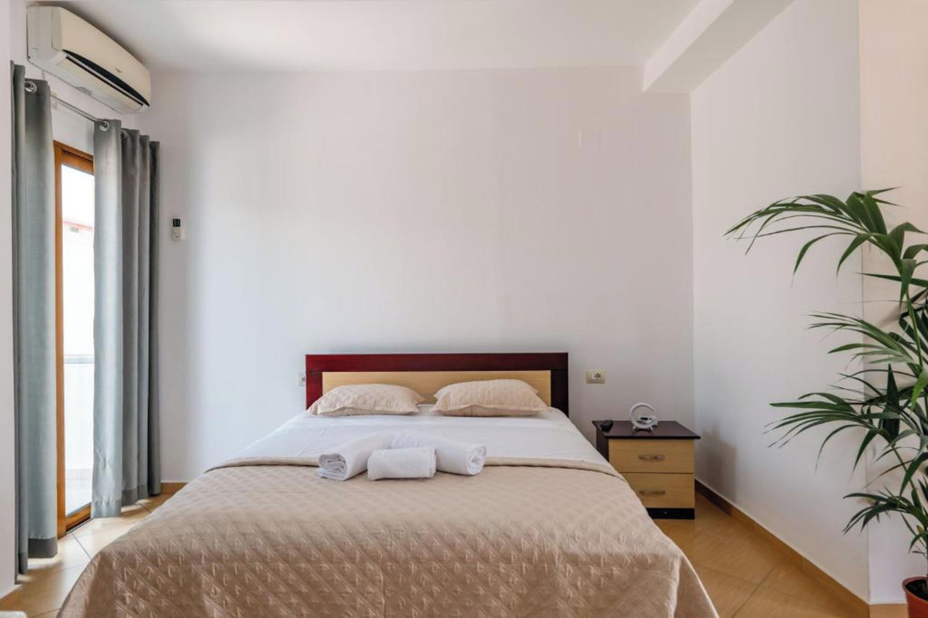 A hotel room with a bed, a bedside table, a potted plant, and an air conditioning unit.