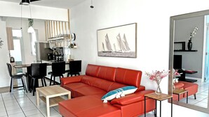 Un salon moderne avec un canapé rouge, un coin repas avec des chaises, une cuisine en arrière-plan et un grand miroir qui reflète la pièce.