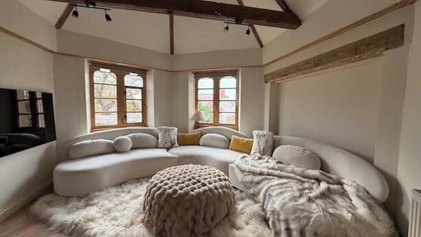 A cozy living room with a curved sofa, a fluffy ottoman, and large windows with wooden frames.