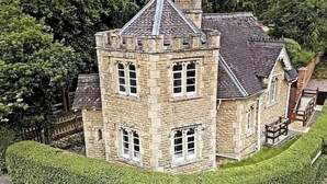 A stone building with a turret, multiple windows, and a well-maintained hedge.