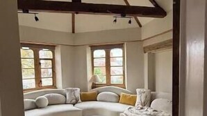A room with a sloped ceiling, a large window, a sofa, an ottoman, and a furry rug.