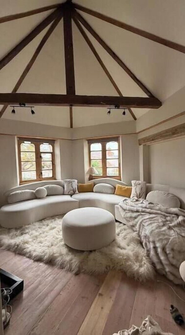 A room with a large sectional sofa, a round ottoman, a fluffy rug, and wooden beams on the ceiling.