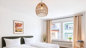A modern bedroom with a large bed, a hanging light fixture, a window with curtains, and a view of buildings outside. - Central living between the Rhine, old town and vineyards (Rüdesheim am Rhein)