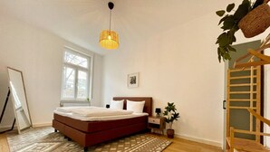 Een slaapkamer met een bed, een raam, een hanglamp, een houten stoel en een bijzettafeltje met een plant.