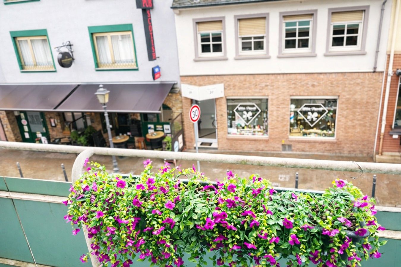 A balcony with hanging flower baskets and a view of a street with buildings and a no parking sign.