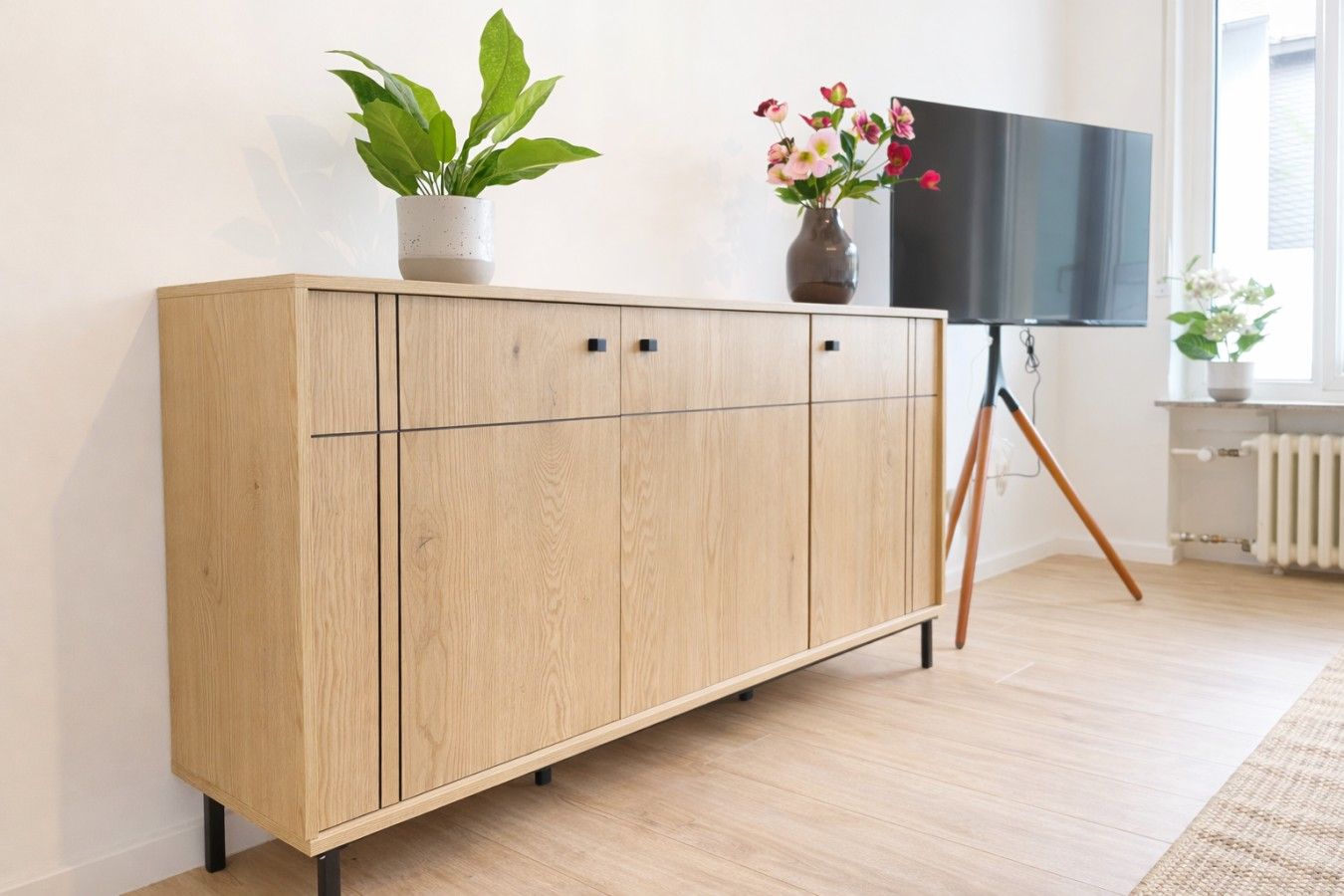 Ein Holzschrank mit einem an einem Gestell befestigten Flachbildfernseher, ein Blumentopf und eine Heizung in einem Raum mit hellem Bodenbelag.
