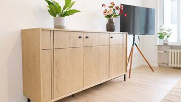 Ein Holzschrank mit einem an einem Gestell befestigten Flachbildfernseher, ein Blumentopf und eine Heizung in einem Raum mit hellem Bodenbelag.