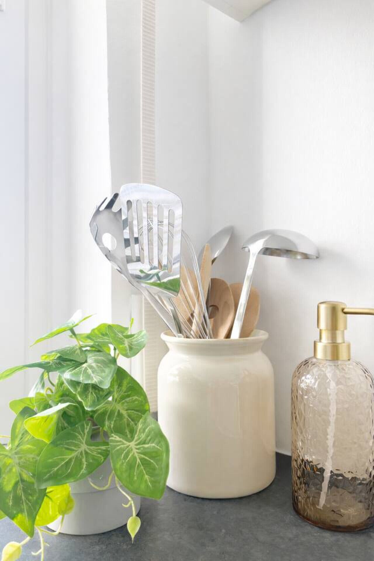 Een aanrecht met een plant, een witte keramieken pot met keukengerei en een glazen zeepdispenser.