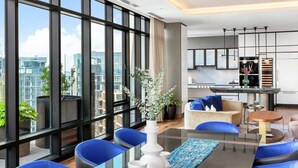 A modern hotel room with large windows, a dining area with blue chairs, a kitchen, and a living area with a sofa and a coffee table. - Luxury Seaport Stay | Rooftop & Pool (Boston, MA)