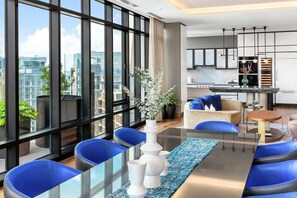 A modern hotel room with large windows, a dining area with blue chairs, a kitchen, and a living area with a sofa and a coffee table. - Luxury Seaport Stay | Rooftop & Pool (Boston, MA)