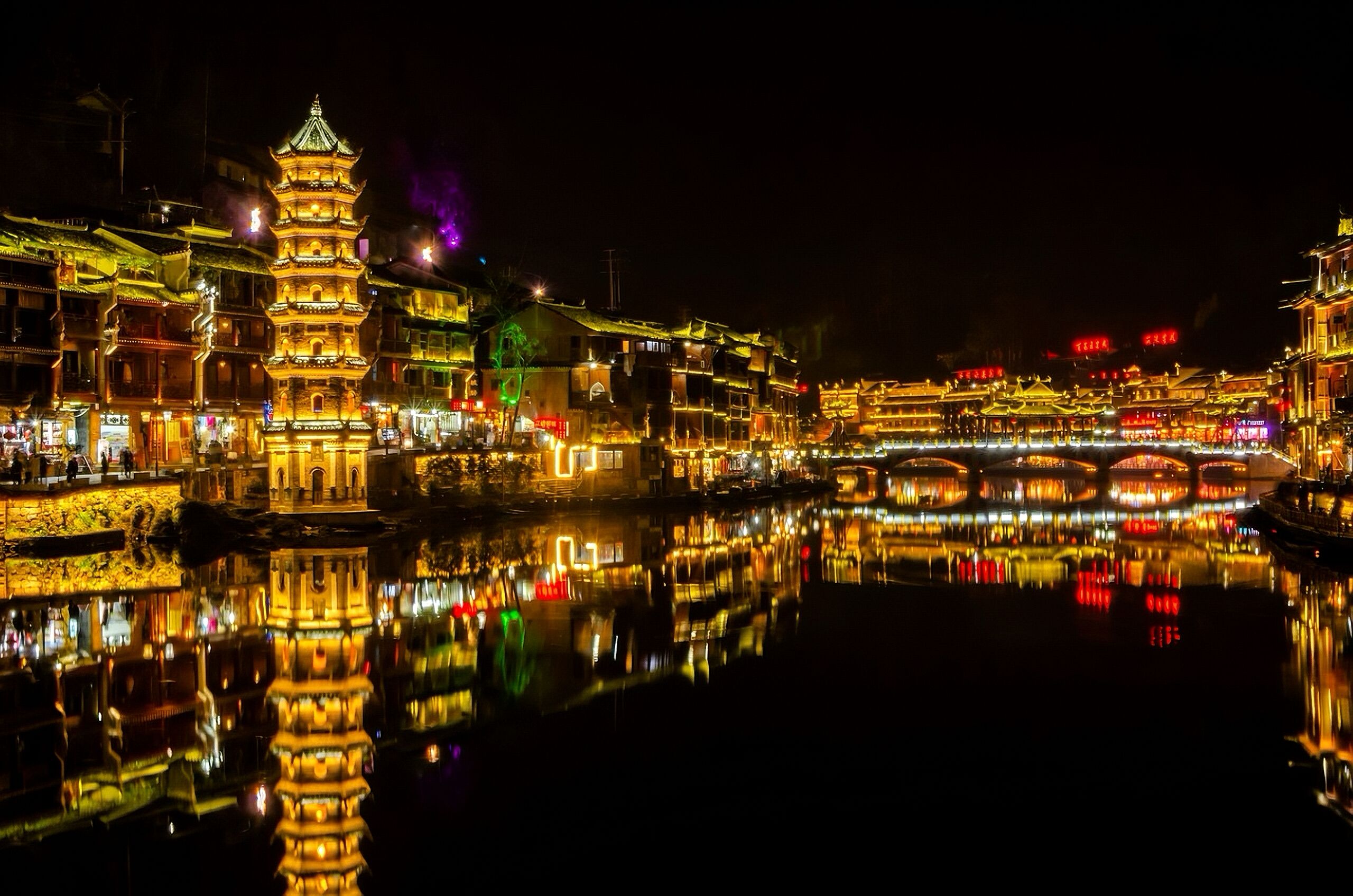 A traditional building with a multi-tiered tower, a reflection in the water, and a bridge in the background.