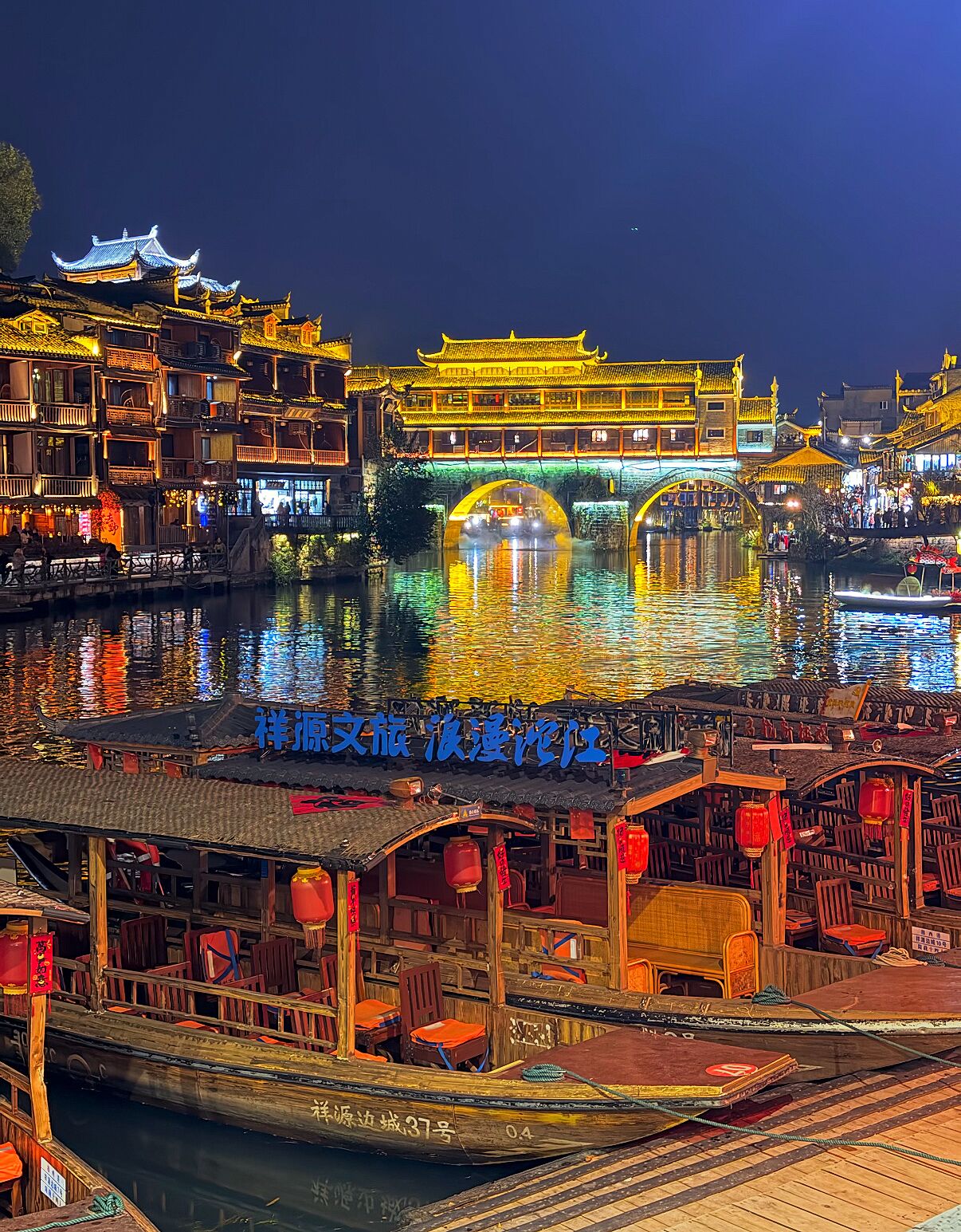 Uma charmosa cidade aquática chinesa à noite, com edifícios iluminados, um barco de madeira atracado em primeiro plano e uma ponte ao fundo.