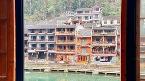 A traditional multi-story building with a wooden balcony, a bridge over water, and a forested hillside in the background. - Fenghuang Meomory Riverview Inn (Xiangxi)