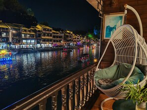 Ein Balkon mit Korbstuhl und Geländer, der auf einen Fluss mit traditionellen Gebäuden und Lichtern blickt.