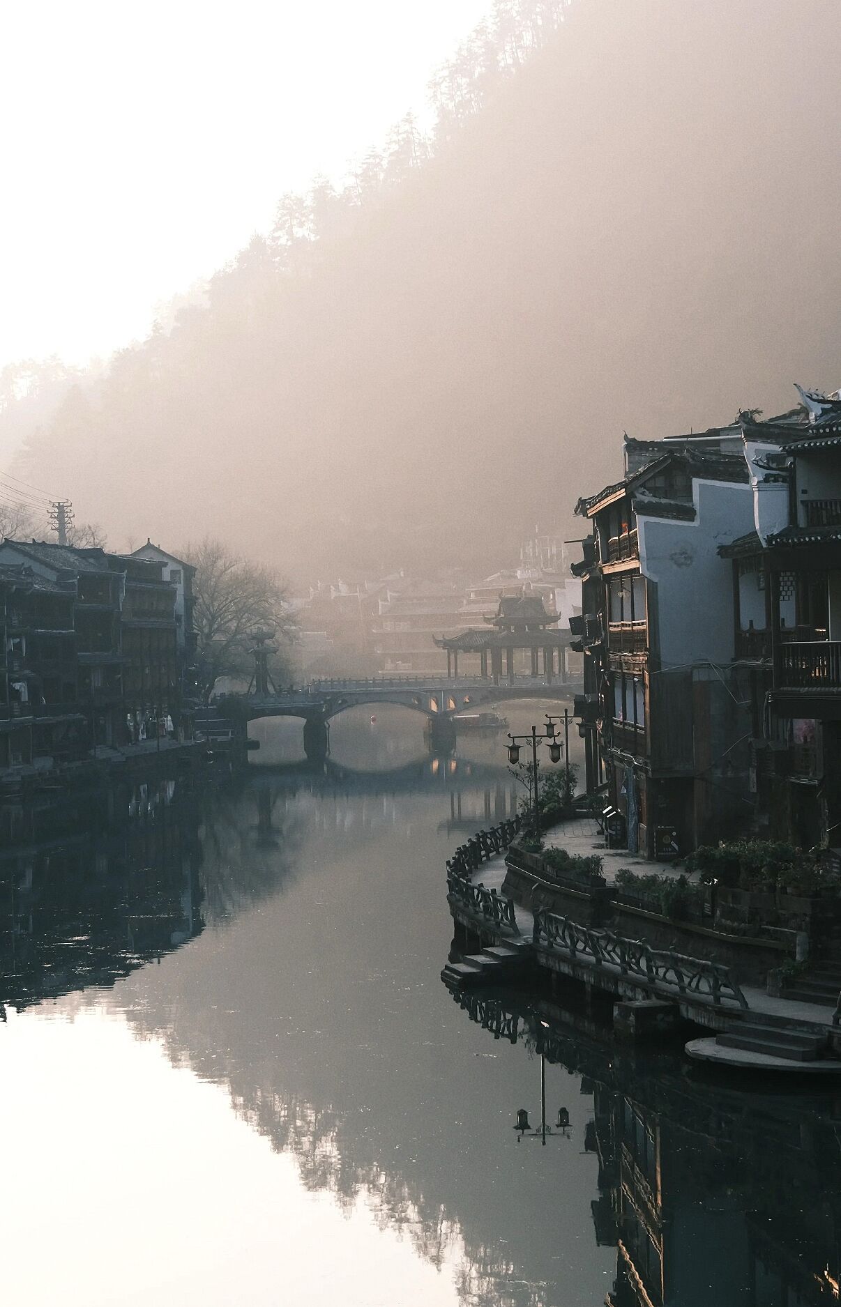 A river with traditional buildings on the banks, a bridge, and a misty atmosphere.