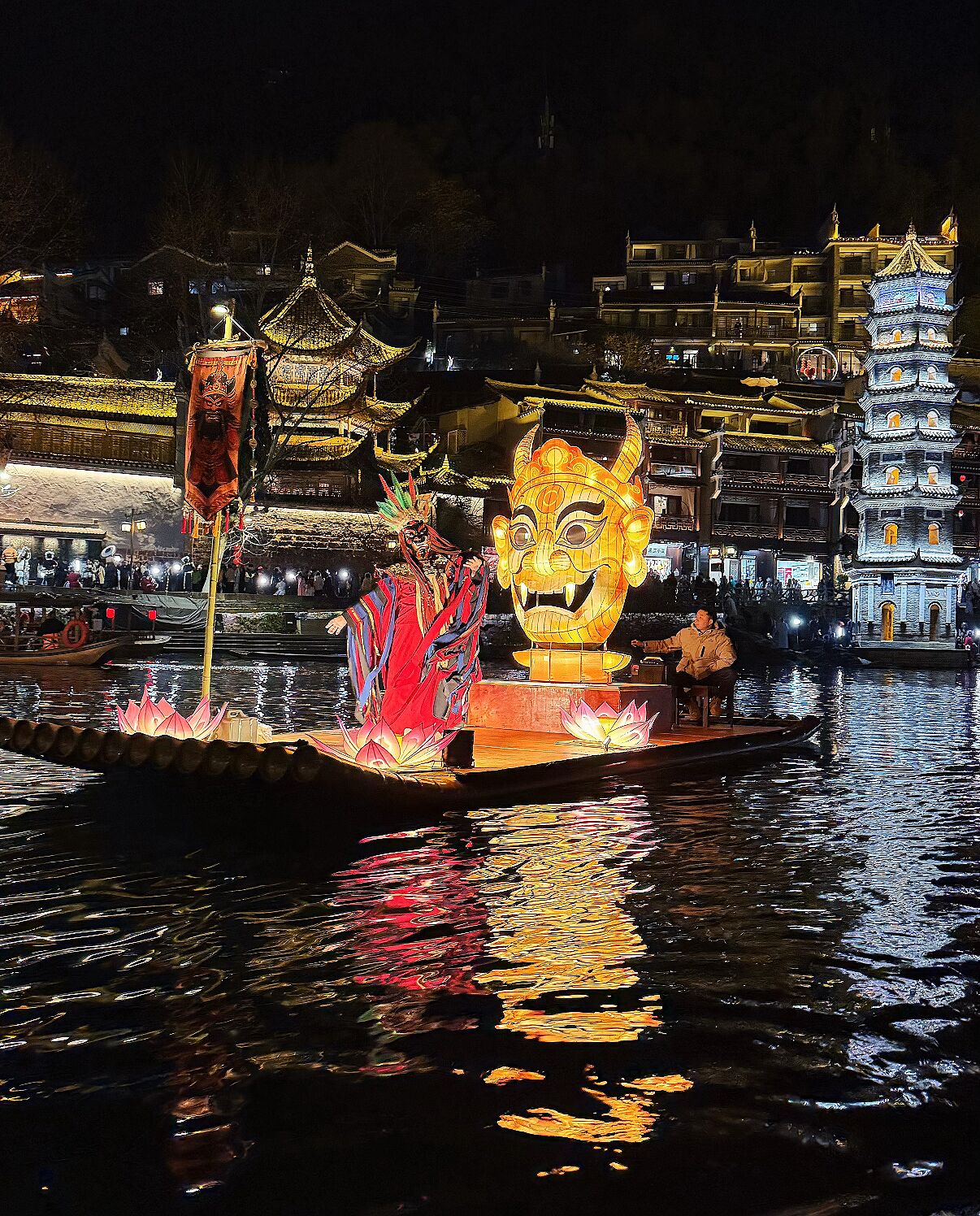 A traditional Chinese water lantern festival with a performer on a boat, illuminated buildings, and a pagoda in the background.