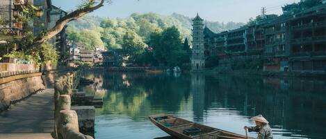 Un barco de madera tradicional en un río tranquilo, un sendero de piedra a la orilla y edificios tradicionales al fondo.
