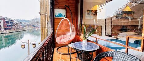 Un balcone con una sedia a dondolo, un tavolino con un vaso e vista su un fiume e degli edifici.