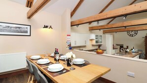 A dining area with a long wooden table set for four, a kitchen in the background, and exposed wooden beams on the ceiling. - Cider Press Cottage (Torpoint)