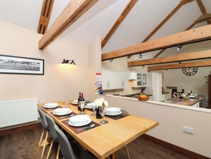 A dining area with a long wooden table set for four, a kitchen in the background, and exposed wooden beams on the ceiling. - Cider Press Cottage (Torpoint)
