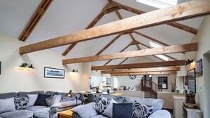 A spacious living room with a high ceiling and exposed wooden beams, a grey sectional sofa set, a small wooden table, and a kitchen in the background. - Cider Press Cottage (Torpoint)