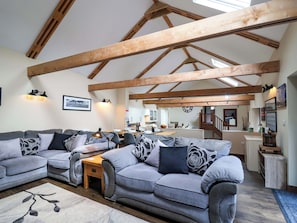 A spacious living room with a high ceiling and exposed wooden beams, a grey sectional sofa set, a small wooden table, and a kitchen in the background. - Cider Press Cottage (Torpoint)