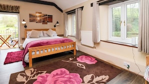 A bedroom with a wooden bed, a large window with curtains, a radiator, and a view of greenery outside. - Cider Press Cottage (Torpoint)